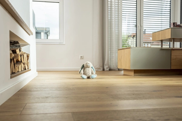 Ein modernes, helles Wohnzimmer mit Holzboden. Im Vordergrund sitzt ein Plüschhase auf dem Boden. Links ist ein Kamin mit gestapeltem Holz zu sehen, rechts ein Holzmöbelstück. Große Fenster mit Jalousien lassen viel Licht herein.