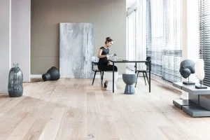 Eine Frau sitzt auf einem Stuhl, an einem Tisch, in ihrem modernen Wohnzimmer. Im Vordergrund steht der helle Parkettboden.