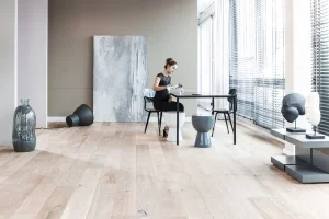 Eine Frau sitzt auf einem Stuhl, an einem Tisch, in ihrem modernen Wohnzimmer. Im Vordergrund steht der helle Parkettboden.