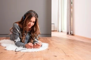 Eine Frau liegt auf dem Bauch auf einem hochwertigen Parkettboden und schaut auf ihr Smartphone. Sie trägt Kopfhörer und unter ihr befindet sich ein weicher, kleiner Teppich.