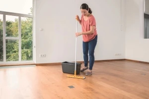 Eine Frau steht in einem großen Zimmer und wischt den Parkettboden mit einem Wischmopp. Neben ihr steht ein Eimer mit Pflegemittel.