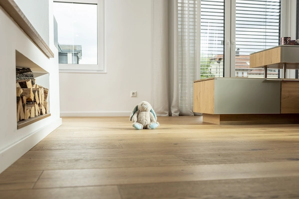Ein modernes, helles Wohnzimmer mit Holzboden. Im Vordergrund sitzt ein Plüschhase auf dem Boden. Links ist ein Kamin mit gestapeltem Holz zu sehen, rechts ein Holzmöbelstück. Große Fenster mit Jalousien lassen viel Licht herein.
